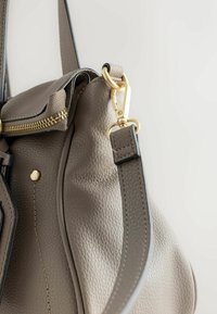 Harmaa nahkainen käsilaukku, jossa on teksturoitu pinta, kullanväriset metalliosat, vetoketjukiinnitys ja irrotettava hihna. Huomionarvoinen yläsauman yksityiskohta.