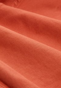 Puuvillakangas vaalean korallinvärisessä sävyssä, jossa on sileä pinta näkyvine ompeluviivoineen ja taittuneita kerroksia, jotka näyttävät pehmeältä laskeutumiselta.