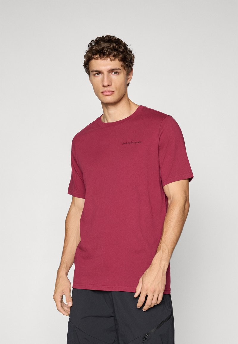 Young man with curly hair wearing a dark red short-sleeve t-shirt and black pants, standing against a plain light background.