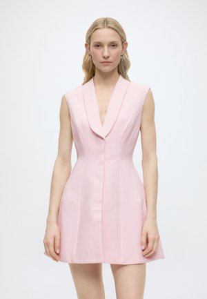 Γυναίκα με μακριά ξανθά μαλλιά, που φοράει αμάνικο, ανοιχτό ροζ ραμμένο φόρεμα με βαθύ V στο ντεκολτέ και μαξιλαρωτούς ώμους, στέκεται μπροστά σε απλό λευκό φόντο.