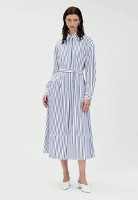 Abito a camicia a righe con maniche lunghe, in tessuto bianco e blu navy, con cintura in vita, gonna plissettata e orlo asimmetrico, abbinato a tacchi bianchi a blocco.