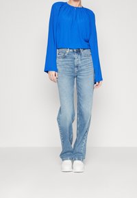 Blusa lunga blu a maniche lunghe con scollo pieghettato abbinata a jeans chiari a zampa d'elefante e sneakers bianche, su uno sfondo neutro.