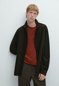 Sort kordfløjlsskjorte over en rød sweater, parret med brune bukser. Skjorten har en knaplukning og en afslappet pasform.