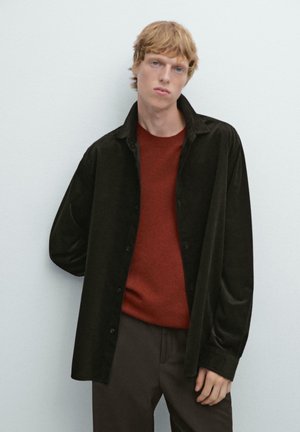 Chemise en velours côtelé noir par-dessus un pull rouge, associée à un pantalon marron. La chemise présente un design à boutons et une coupe décontractée.