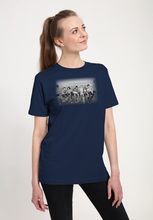 Femme avec une longue queue de cheval portant un t-shirt bleu marine avec une photo en noir et blanc de cinq ouvriers assis sur une poutre au-dessus d'une ville.