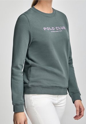 Femme portant un sweat-shirt vert foncé avec un texte "POLO CLUB Established 2012" en rose et blanc, associé à un pantalon blanc.