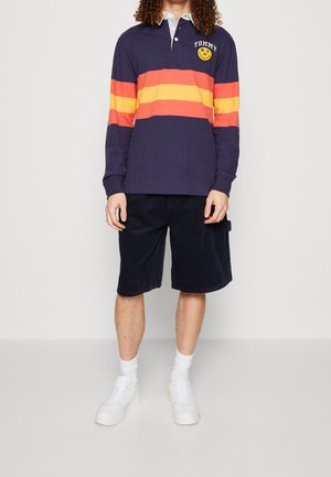 Polo à manches longues bleu marine avec des rayures horizontales rouges et oranges, un col blanc et un logo souriant ; associé à un short en velours côtelé noir.