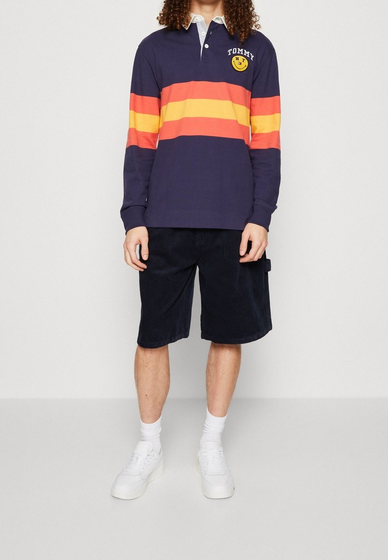 Polo de manga larga en color azul marino con rayas horizontales rojas y naranjas, cuello blanco y un logo sonriente; acompañado de pantalones cortos de pana oscuros.