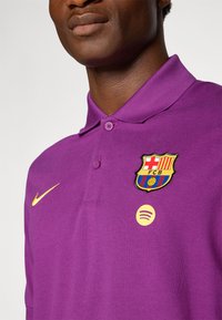 Paarse poloshirt met een kraag, voorzien van een geel Nike-logo, geborduurd FC Barcelona-embleem en een ronde Spotify-logo op de borst.