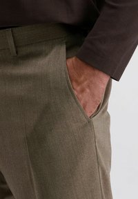 Man in een bruine visgraatjas met zijn hand in de zak van olijfgroene broek tegen een effen achtergrond.