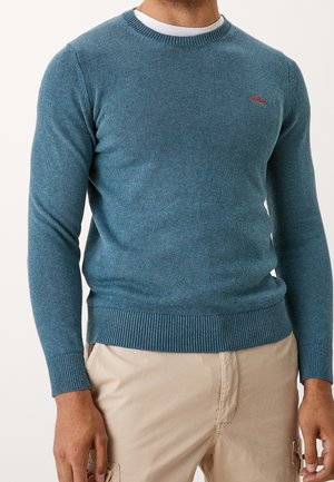 Homme portant un pull col rond bleu sarcelle par-dessus un sous-vêtement blanc avec un pantalon cargo beige, debout, les bras détendus le long du corps.