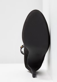 Chaussure à talons hauts en suede noir avec un bout pointu et un talon fin. Dotée d'une bride à la cheville à motif floral avec des accents multicolores.