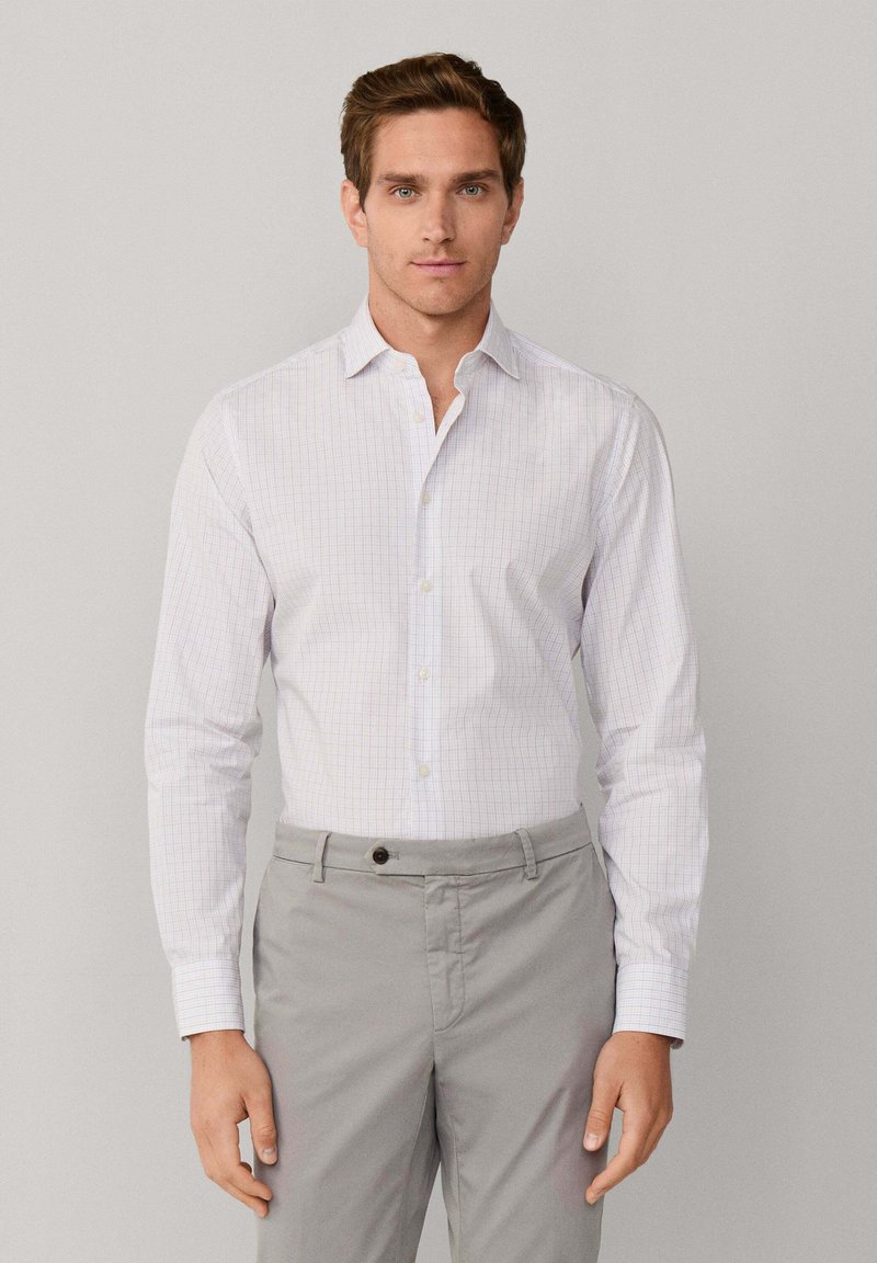 Camicia bianca a maniche lunghe con un sottile motivo a griglia blu e rosso, dotata di colletto classico e chiusura con bottoni, abbinata a pantaloni grigi.