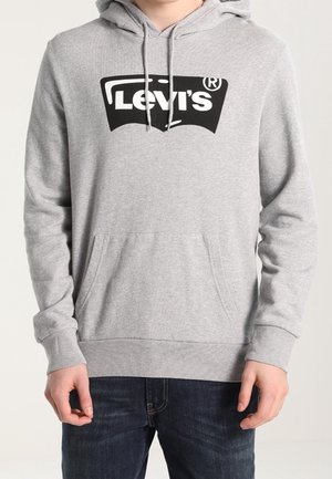 Persona vistiendo una sudadera ligera de Levi's en gris claro con un logo negro, con un bolsillo canguro frontal y capucha con cordones, combinada con unos jeans azul oscuro.