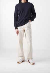 Marinblå stickad tröja med rund hals och grön logotyp, kombinerad med krämfärgade vida byxor och vita sneakers med lila och orange detaljer.