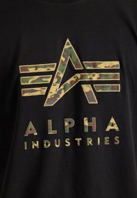 Juodas marškinėliai su kamufliažo grafikų dizainu, kuriame yra abstrakti raidė "A" ir tekstas "ALPHA INDUSTRIES" panašiomis spalvomis.