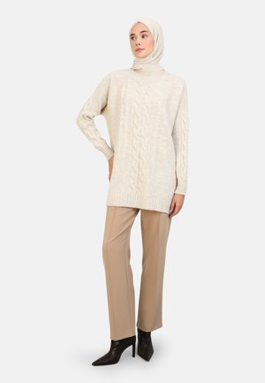 Knit sweater in light beige with a cable pattern, featuring a high neckline and ribbed cuffs, paired with beige trousers and black heels.