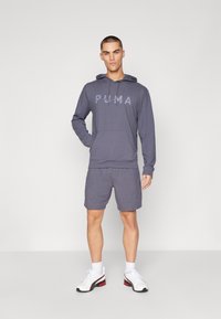 Grauer Hoodie mit frontalem Taschen- und Kordelzug, versehen mit einem strukturierten PUMA-Logo. Kombiniert mit grauen Shorts und weißen Sportschuhen.