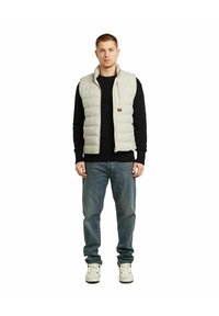 Gilet imbottito grigio chiaro con colletto alto, chiusura con zip e logo ricamato. Indossato sopra un maglione nero a coste e abbinato a jeans blu e scarpe da ginnastica bianche.