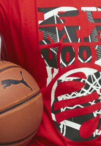 Červené bavlněné tričko s výrazným černým grafickým textem a designem basketbalové sítě, doplněné hnědým basketbalem s černým logem.