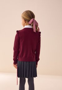 Bordeaux gebreide trui met ruches, gecombineerd met een geplooide grijze rok. Haar opgestoken met een roze ruitlint, en ze houdt een roze voorwerp vast.