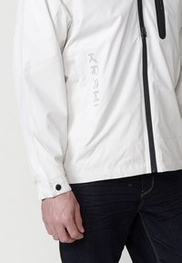 Witte waterdichte jas met een rits aan de voorkant, zwarte accenten en een knoopmanchet; heeft een bedrukte logo aan de zijkant.