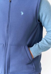 Gilet blu con zip realizzato in tessuto morbido con due tasche frontali, caratterizzato da dettagli in maglia di colore blu più chiaro sulle maniche e logo ricamato sul petto.