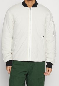 Padded bomber jacket in off-white with a black collar, zippered front, side pockets, and a black logo accent on the chest.
