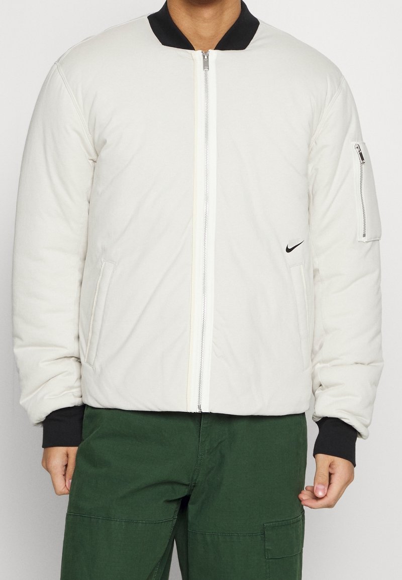 Padded bomber jacket in off-white with a black collar, zippered front, side pockets, and a black logo accent on the chest.