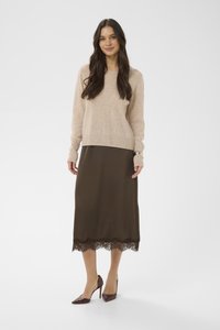 Femme debout portant un pull beige à manches longues, une jupe midi marron avec une bordure en dentelle, et des chaussures à talons marron sur un fond blanc.