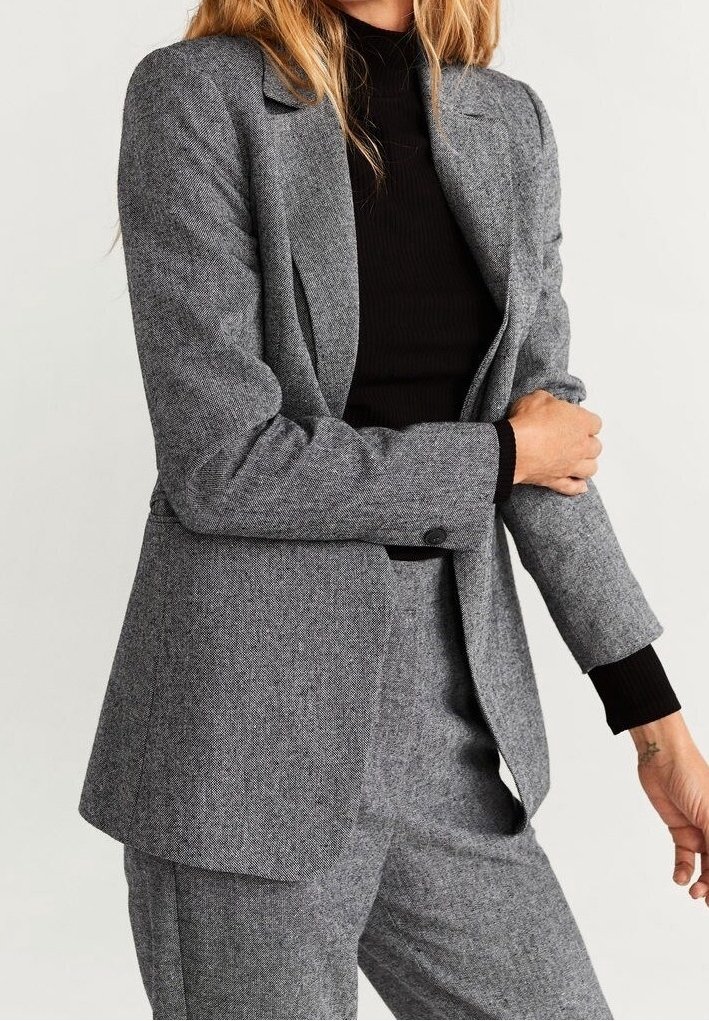 Femme portant un costume gris texturé avec un blazer cintré et un pantalon assorti, associé à un pull à col roulé côtelé noir.