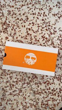 Biały pudełko na buty z pomarańczowym poziomym pasem, na którym widnieje białe logo stylizowanych korzeni drzewa; umieszczone na nakrapianej podłodze.