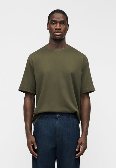 Homme à la peau foncée portant un T-shirt à manches courtes vert olive et un pantalon bleu foncé, debout devant un fond clair uni.