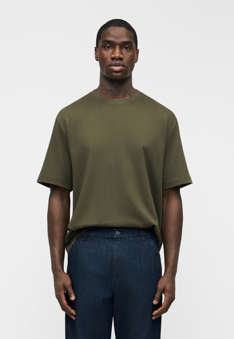 Ανδρας με σκουρόχρωμο δέρμα που φοράει λαδί κοντομάνικο μπλουζάκι και σκούρο μπλε παντελόνι, στέκεται μπροστά σε απλό ανοιχτό φόντο.