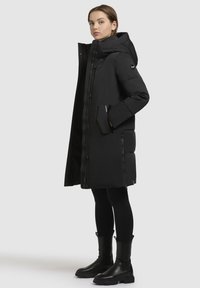 Frau in einem langen, gepolsterten schwarzen Mantel mit Kapuze, schwarzi Leggings und schwarzen Stiefeletten, steht vor einem einfarbigen hellgrauen Hintergrund.