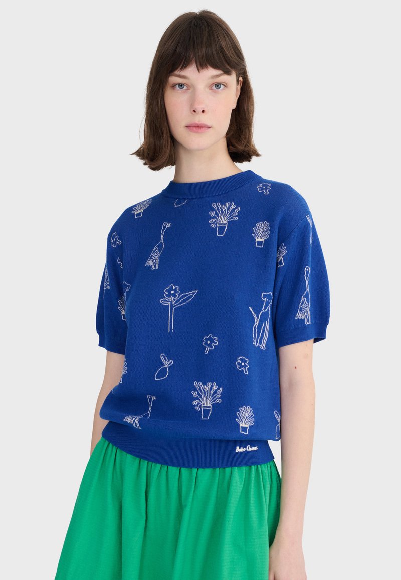 Kobieta z krótkimi brązowymi włosami, ubrana w niebieski sweter z krótkim rękawem ozdobiony białymi wzorami roślinnymi i zwierzęcymi oraz jasnocyemnozieloną spódnicę.