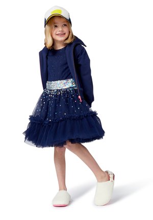Ein junges Mädchen trägt ein marineblaues Pailletten-Kleid, eine marineblaue Jacke, eine weiße Mütze und weiße, flauschige Slipper. Sie steht mit einem Bein leicht angehoben.