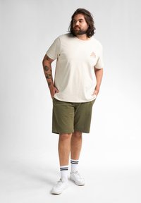 Beige katoenen t-shirt met een klein oranje logo, gecombineerd met olijfgroene shorts. Witte sneakers met zwartgestreepte sokken. Eenvoudig ontwerp, casual pasvorm.