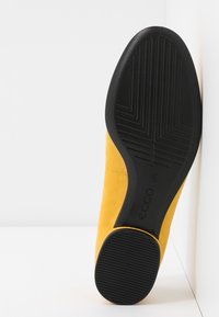 Chaussure en daim jaune vue du dessous, montrant une semelle extérieure en caoutchouc noir avec des motifs de sculpture et un petit logo près de l'arche.