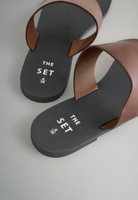 Pruuni nahast libisevad sandaalid musta kummist tallaga, millel on avatud disain ja valge trükitud tekst: "THE SET 6 39". Sile tekstuur.