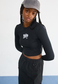Fekete hosszú ujjú cropped top, ami fehér Nike logóval rendelkezik, fekete cargo nadrággal párosítva. Szürke sapka kontrasztot ad az öltözethez.