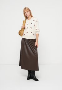 Cardigan crème avec broderie florale, associé à une jupe longue en simili cuir marron. Des bottes noires montantes complètent la tenue.