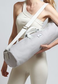 Bolsa de yoga cilíndrica gris, hecha de una tela resistente, con una correa para el hombro y asas de transporte, y una pequeña etiqueta con el logotipo.