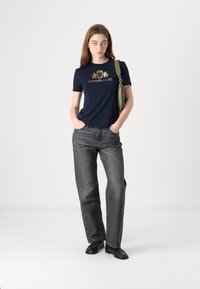 T-shirt marine avec un logo doré graphique, associé à un jean gris à jambes larges. Le modèle tient un sac à main vert. Des chaussures noires complètent le look.