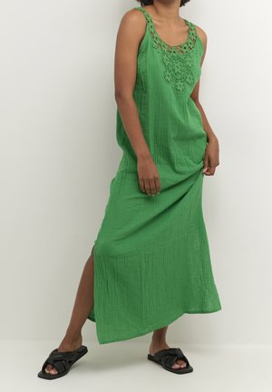 Woman wearing a sleeveless green maxi dress with crochet neckline and black slip-on sandals, standing against a plain white background.
