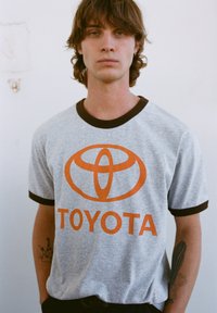 Sivé tričko s hnedými akcentmi, ozdobené oranžovým logom Toyota a textom. Vyrobené z bavlny s klasickým okrúhlym výstrihom.