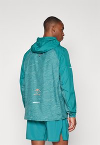 Turquoise windbreaker met een gestructureerde achterkant, verstelbare capuchon, reflecterende accenten en logo-detail. Gepaard met bijpassende turquoise shorts.