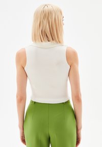 Top bianco a coste senza maniche con colletto, abbinato a pantaloni verdi a vita alta. La vista posteriore mostra linee pulite e una vestibilità strutturata.