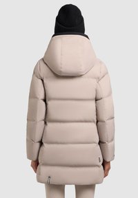 Jacheta puffer de culoare bej deschis, cu glugă, cu un design matlasat și o croială ușor mai lungă. Include o caciulă tricotată neagră.