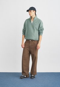 Vaaleanvihreä vetoketjullinen huppari, tummanruskeat leveät housut, musta lippis ja valkoisilla yksityiskohdilla varustetut kengät. Silkkinen kangas, rento malli ja vapaa-ajan tyyli.